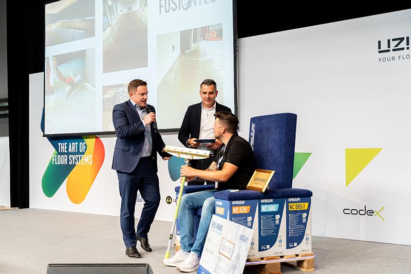 Op het podium van een vloeren vakbeurs spreekt een man in pak in een microfoon, terwijl een andere man, zittend op een stoel die is gemaakt van opgestapelde zakken met egalisatie, een rakel vasthoudt.