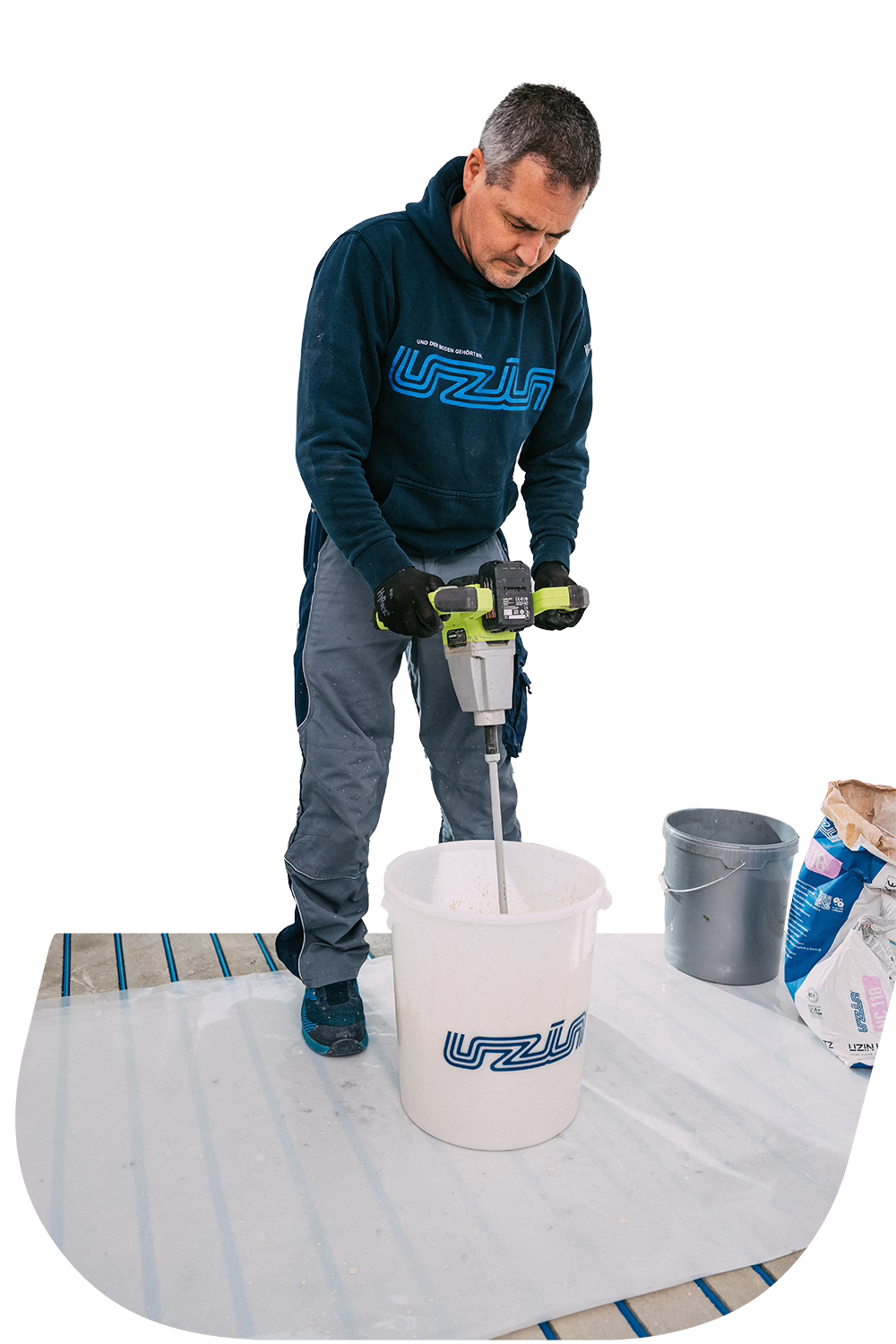 A man in a navy hoodie and gray pants uses a power mixer to stir material in a large white bucket. Nearby are a gray bucket and a bag of ingredients on a plastic-covered floor.