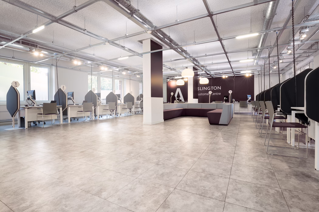 UZIN_Application Image_KE 66_Inslington Customer Care Centre_GB_2020-06 A bright, modern customer service hall with a wide open floor and light grey tiled flooring. Along the left and right walls are rows of white service desks with high-backed dark privacy panels and numbered signs above each workstation; each desk has a computer monitor and a simple visitor chair. The center of the room features a large square column and a modular purple-and-grey seating area with pendant lamps hanging above. The ceiling shows an exposed structural grid with suspended lighting rails and visible ventilation ducts, giving a contemporary industrial look. Large windows along the left wall admit natural daylight; the space is clean, minimal and currently unoccupied.