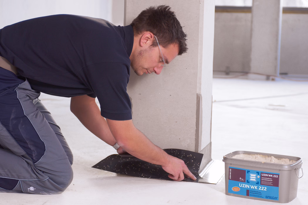 Een vakman met veiligheidsbril en werkkleding knielt in een bijna lege binnenruimte en drukt een zwarte lijm- of onderlegdeken bij de onderkant van een lichte muur of kolom aan. Met zijn handen strijkt hij het materiaal langs de vloerafwerking glad. Dichtbij staat een kunststof bak met het opschrift 'UZIN WK 222' met lijm erin. De ruimte is schoon en onafgewerkt met een beton- of dekvloer en weinig achtergronddetails.