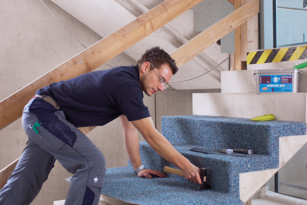 Een vakman legt een blauwe, gerecyclede vloerbedekking op betonnen traptreden in een onafgewerkt gebouwinterieur. Hij knielt en leunt naar voren, draagt een veiligheidsbril, een donkerblauw poloshirt en grijze werkbroeken, en gebruikt een rubberen hamer om het tapijt op de treden aan te drukken; in de buurt liggen een troffel, een rolmaat en een plastic bak met het opschrift 'UZIN WK 222'. De scène is goed verlicht met natuurlijk licht, tijdelijke houten leuningen zijn zichtbaar, de focus ligt op de zorgvuldige installatie en de textuur van het tapijt op de treden.